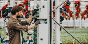 A man operates a machine inside a greenhouse, surrounded by plants and natural light.