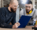 Two men in work clothes examine a blue folder, discussing its contents with focused expressions.
