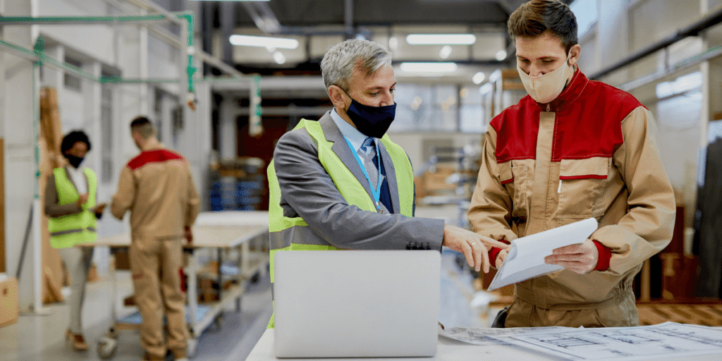 Two men in work clothes and masks collaborate on a laptop, focused on their task in a professional setting.