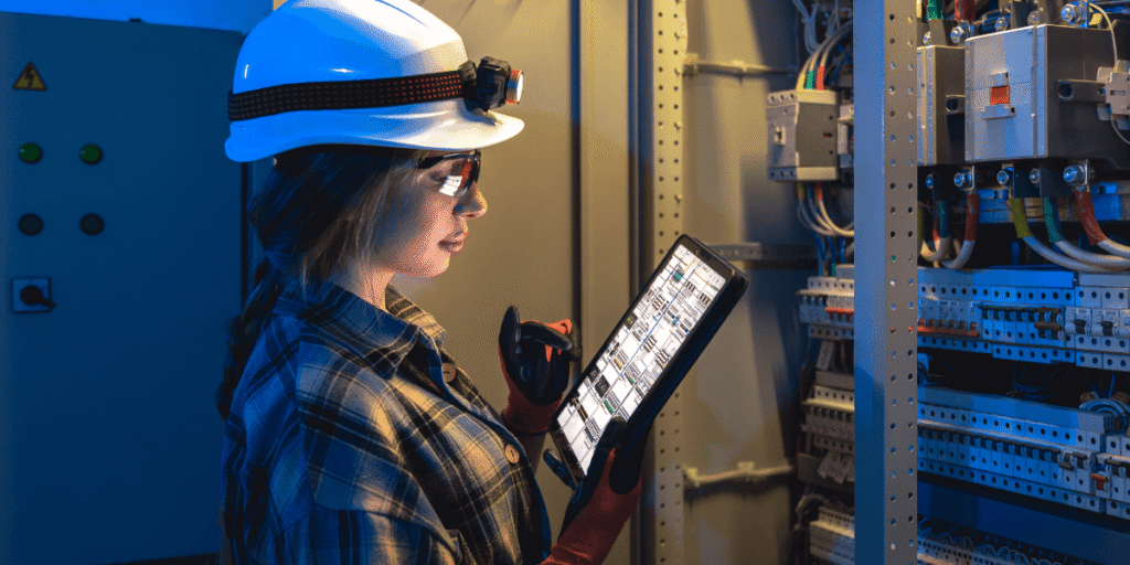 A woman wearing a hard hat and helmet, holding a tablet, appears focused on her work in a construction environment.