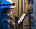 A woman wearing a hard hat and helmet, holding a tablet, appears focused on her work in a construction environment.