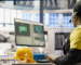 A woman wearing a yellow shirt and hard hat is focused on her work at a computer.
