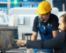 Two men wearing blue hard hats collaborate on a computer, focused on their work in a construction or engineering setting.
