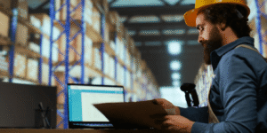 A man wearing a hard hat sits at a table, focused on his laptop, indicating a work-related environment.